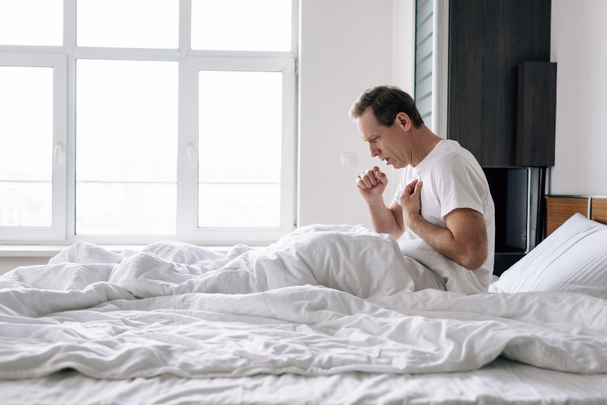 Mann sitzt hustend im Bett - Trockener Hals